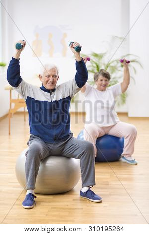 Elderly Man And Woman Exercising On Gymnastic Balls During Physiotherapy Session At Hospital