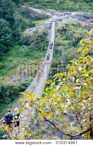 A Group Of Trekkers Crossing The Bridge In Valley With River On Manaslu Circuit With View Of Mount M