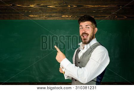 Teacher In Classroom Near Blackboard Pointing At Copy Space. Male Teacher With Pencil In Classroom. 