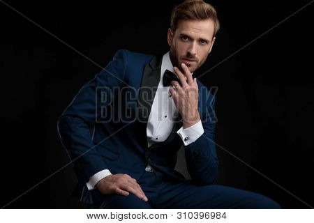 Thoughtful young man holding his hand on his chin while sitting and leaning on his leg, wearing an elegant blue tuxedo on black studio background