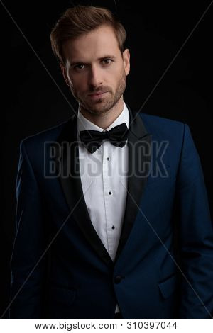 Serious looking groom posing while wearing a blue tuxedo and a black bowtie, standing and posing on black studio background