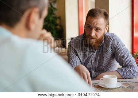 Young Bearded Man Explain Something To His Interlocutor Friend Sitting Indoors Cafe With Cup Of Coff