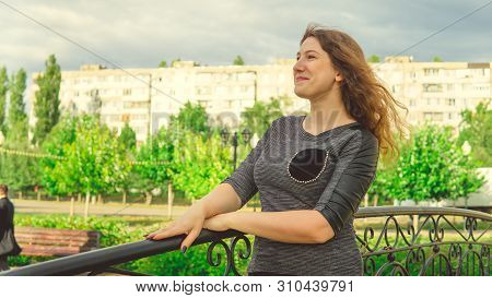 Content Woman Looking Dreamily Standing In Green Park Against Ornamental Iron Fence. Shy Woman Leani