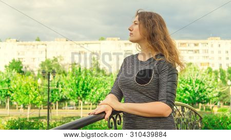 Content Woman Looking Dreamily Standing In Green Park Against Ornamental Iron Fence. Shy Woman Leani