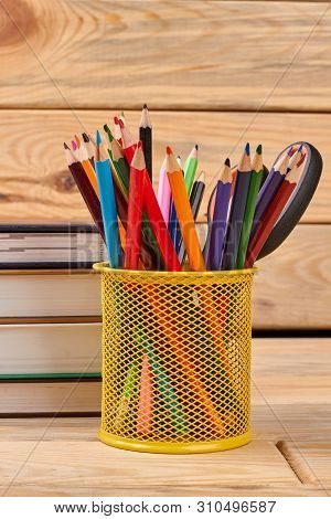 Metal Holder With Multicolored Pencils. Stack Of Books And Colorful Pencils In Basket On Wooden Back