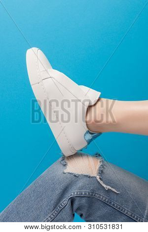 Trendy White Eco Leather Sheakers On Bent Leg Knee In Ragged Jeans Over Pastel Turquoise Blue Backgr