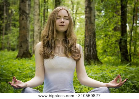 A Girl Is Meditating In Nature. A Blonde Meditates In The Forest In A Lotus Position.