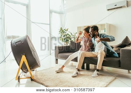 Excited African American Man And Woman Sitting On Sofa In Front Of Blowing Electric Fan