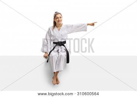 Full length portrait of a young female in kimono and black belt sitting on a panel and pointing to the side isolated on white background