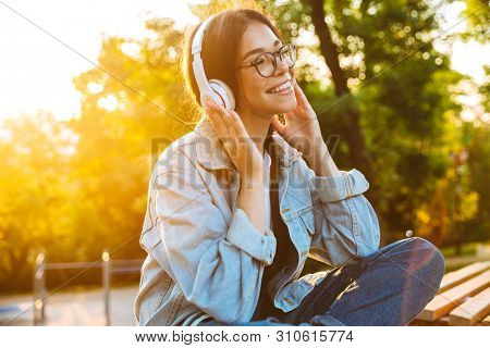 Image of a optimistic smiling young teenage girl student sitting outdoors in beautiful green park listening music with headphones.