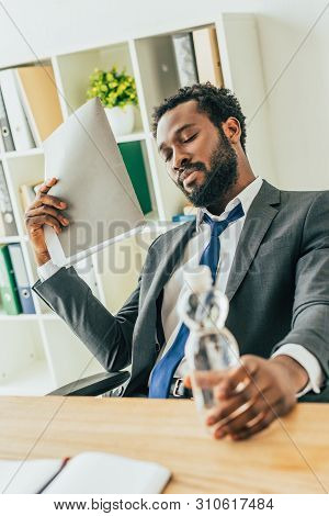 Exhausted African American Businessman Holding Bottle Of Water And Folder While Suffering From Heat 