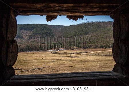 View From Big Window To The Field With A Farm And A River. A Large Frame Of Logs. Against The Backgr