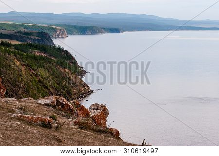 The Rocky Shore Of Lake Baikal With Protruding Stones And Red Moss On Them. The Shore Is Overgrown W