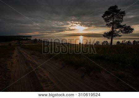 Sunset On The Lake With A Tree And A Sandy Road. The Sun Shines Through The Clouds. Solar Path On Th