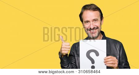 Handsome middle age man holding paper with question mark happy with big smile doing ok sign, thumb up with fingers, excellent sign