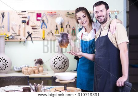 Happy Cacucasian Couple Of Potters In Workshop Posing With Clay Jars Against Instruments Board. Hori