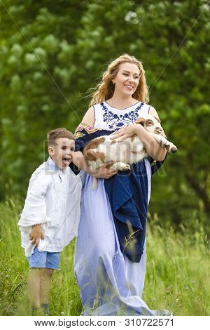Portrait Of Smiling Mother With Her Little Son. Holding Cub Of Newborn Lamb Outdoors On Nature Backg