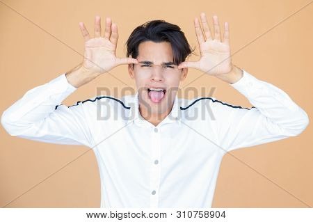 Grimacing Guy Sticking Tongue Out And Holding Hands Near Temples. Portrait Of Young Man In White Shi