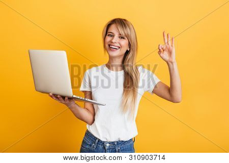 Image of a pleased optimistic emotional young blonde woman posing isolated over yellow wall background dressed in white casual t-shirt using laptop computer showing okay gesture.