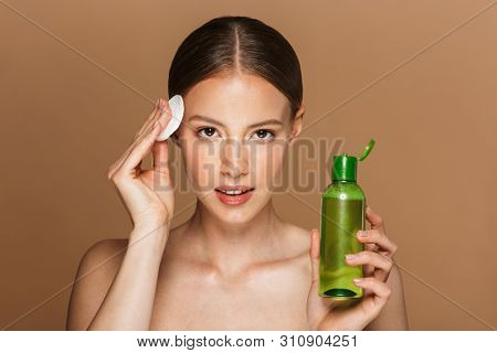 Beauty portrait of an attractive smiling blonde topless woman standing isolated over beige background, cleancing her face with tonic and cotton pad