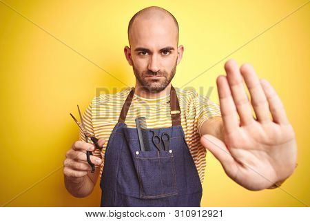Young hairdresser man wearing barber apron holding scissors over isolated yellow background with open hand doing stop sign with serious and confident expression, defense gesture