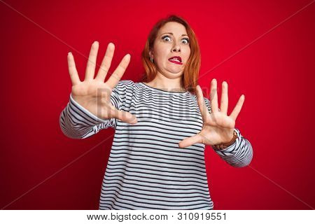 Young redhead woman wearing strapes navy shirt standing over red isolated background afraid and terrified with fear expression stop gesture with hands, shouting in shock. Panic concept.