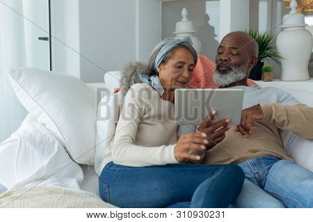 Front view of happy diverse senior couple using a digital tablet on sofa in beach house. Authentic Senior Retired Life Concept