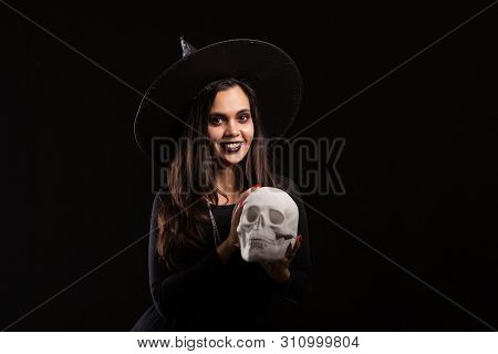 Malefic Witch Woman With A Big Hat Looking Into The Camera With A Human Skull In Her Hands
