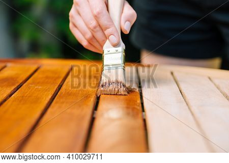 Hand Holding A Brush Applying Varnish Paint On A Wooden Garden Table - Painting And Caring For Wood 