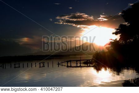 Next To The Pier, The Sun Over The Mountains, A Sundog And Clouds Are Reflected In The Calm Lake In 