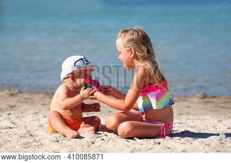 Sister Feeds Her Brother On The Beach With Watermelon. Girl And Little Boy Are Eating On The Beach. 