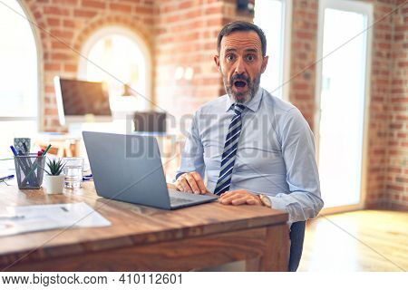 Middle age handsome businessman wearing tie sitting using laptop at the office afraid and shocked with surprise expression, fear and excited face.
