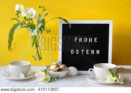 Text Frohe Ostern Means Happy Easter. Text On Letter Board With Freesia Flowers On Table. Black Lett
