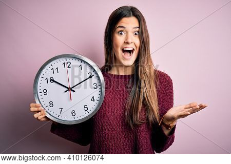 Young beautiful girl doing countdown holding big clock over isolated pink background very happy and excited, winner expression celebrating victory screaming with big smile and raised hands