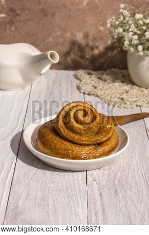 Freshly Baked Cinnamon Bun Stuffed With Spices And Cocoa On A White Wooden Table With A Tough Shadow