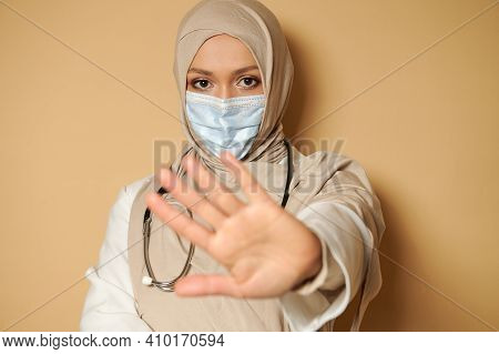 Portrait Of Muslim Doctor Woman In Hijab And Medical Mask Gesturing Stop With Her Hand. Beige Backgr