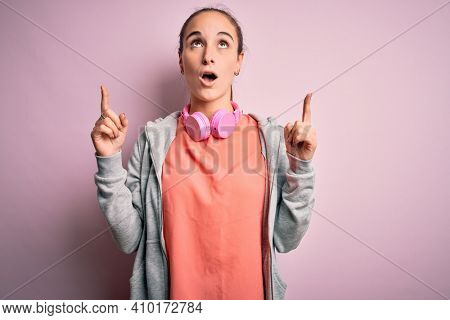 Beautiful sporty woman doing sport listening to music using headphones over pink background amazed and surprised looking up and pointing with fingers and raised arms.