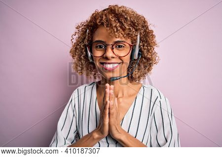 African american curly call center agent woman working using headset over pink background praying with hands together asking for forgiveness smiling confident.