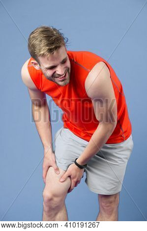 Sportsman With Knee Pain On Violet Background. Man In Orange Vest And Blue Shorts. Health Problem An