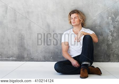 Young handsome man model wearing jeans and white thirt sitting on the floor. Studio fashion portrait. Lifestyle and people concept.