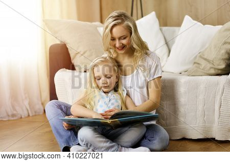 Happy Family. Blonde Young Mother Reading A Book To Her Cute Daughter While Sitting At Wooden Floor 