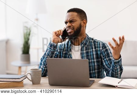 Displeased Black Business Guy Talking On Phone Having Quarrel By Cellphone Sitting At Workplace In M