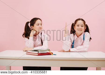 Shoollife Of Twin Schoolgirls Sitting Behind Schooldesk. Frontal View. Over Pink
