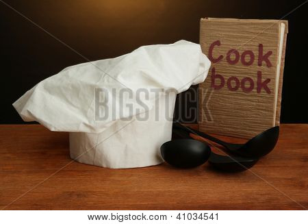 Chef's hat med skedar och kokbok på bordet på brun bakgrund