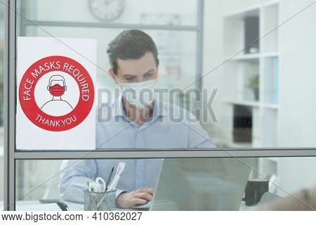Announcement about requirement of wearing protective mask on glass wall of modern office with young businessman networking behiind