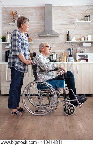 Wife Pushing Invalid Senior Dreamy Man In Wheelchair In Kitchen, Discussing And Smiling, Enjoying Th