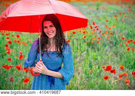 Young beautiful smiling woman wearing summer drees holding red umbrella in poppy field. Charming girl walks before the rain, nature outdoor
