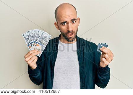 Young hispanic man playing poker holding dollars banknotes and chips in shock face, looking skeptical and sarcastic, surprised with open mouth 