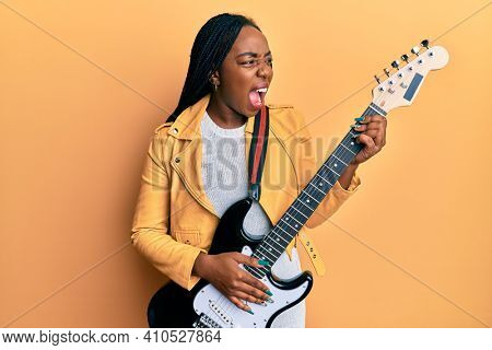 Young african american woman playing electric guitar angry and mad screaming frustrated and furious, shouting with anger. rage and aggressive concept. 