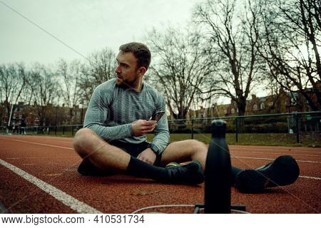 Caucasian Male Athlete Sitting On Trakc Field With A Bottle Of Water Typign On Cellulae Device